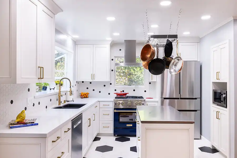 White wood cabinets with brass fixtures, black and white geometric tile flooring, stainless steel appliances with cobalt blue gas stove/oven. Island in center with pot rack above