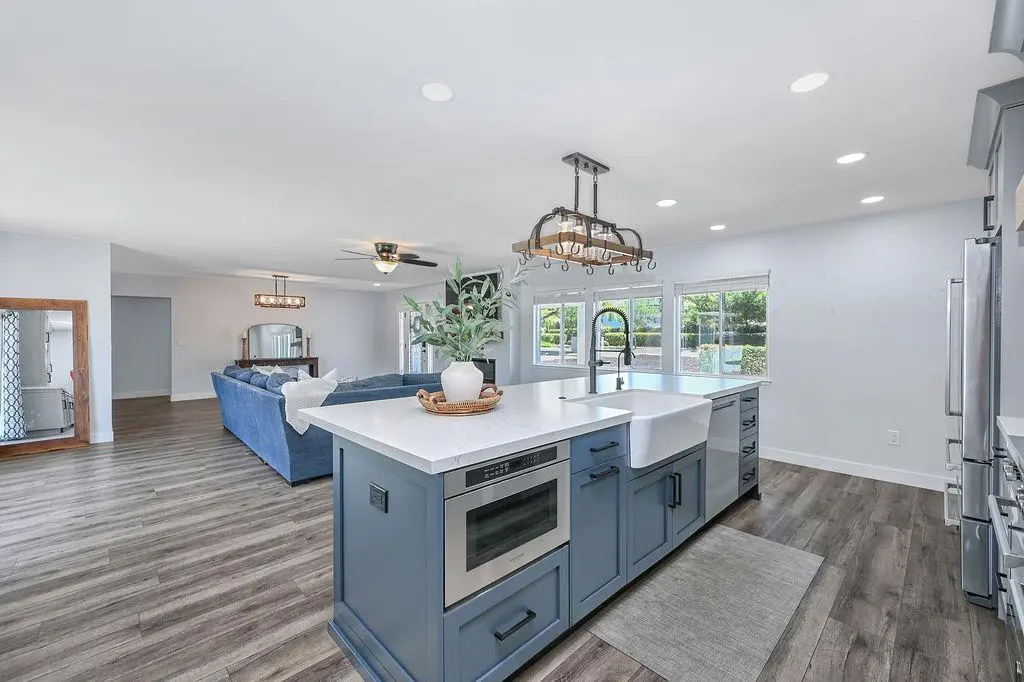 Blue painted island with Signature microwave drawer, Miele dishwasher, and farmhouse sink under a bronze pot-rack chandelier, open to the family room.