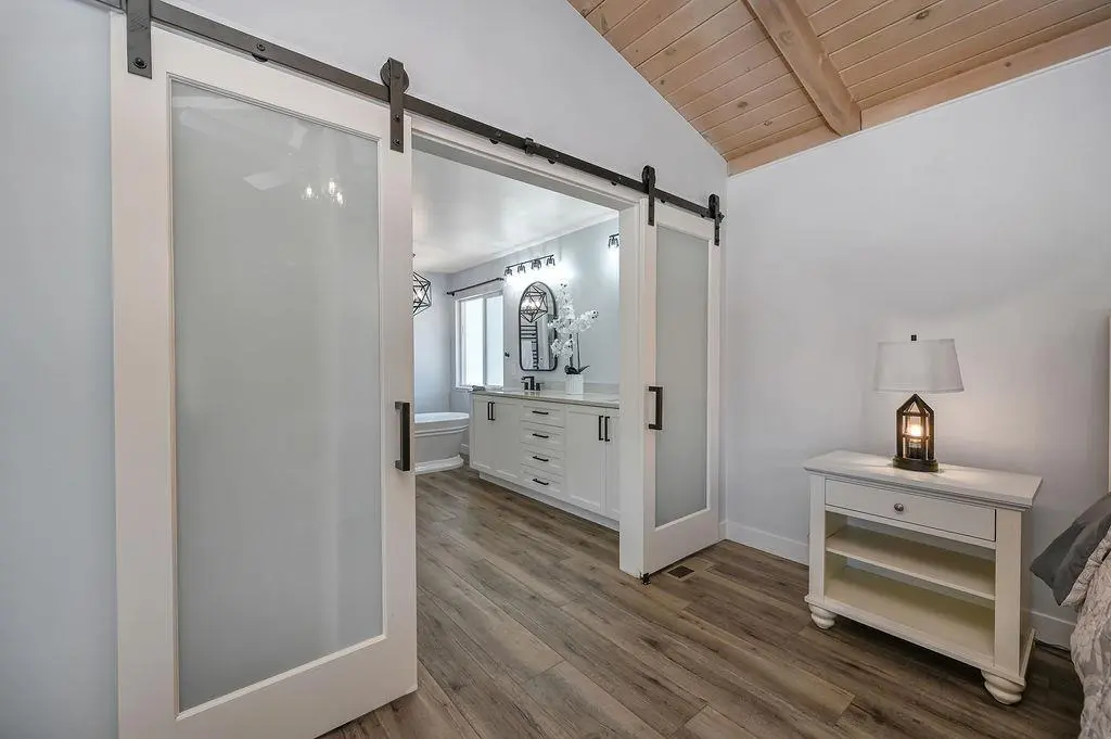 Satin-glass barn doors on matte-black hardware opening from the primary bedroom to a white double vanity and freestanding tub.