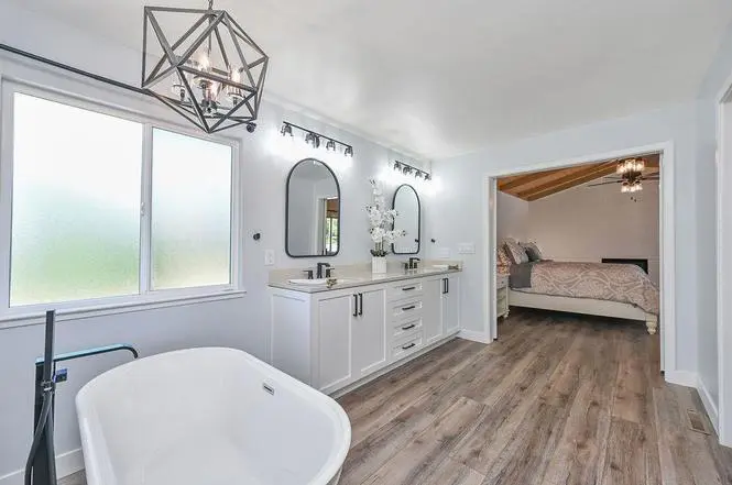 Double vanity with Kohler Caxton sinks and black Moen Genta faucets, a floor-mounted tub filler, and a view back to the primary bedroom.